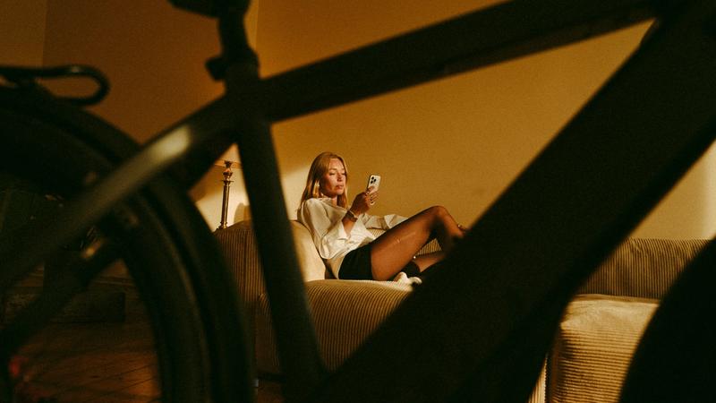 A woman sits on a couch, looking at her phone, with a bicycle frame in the foreground.